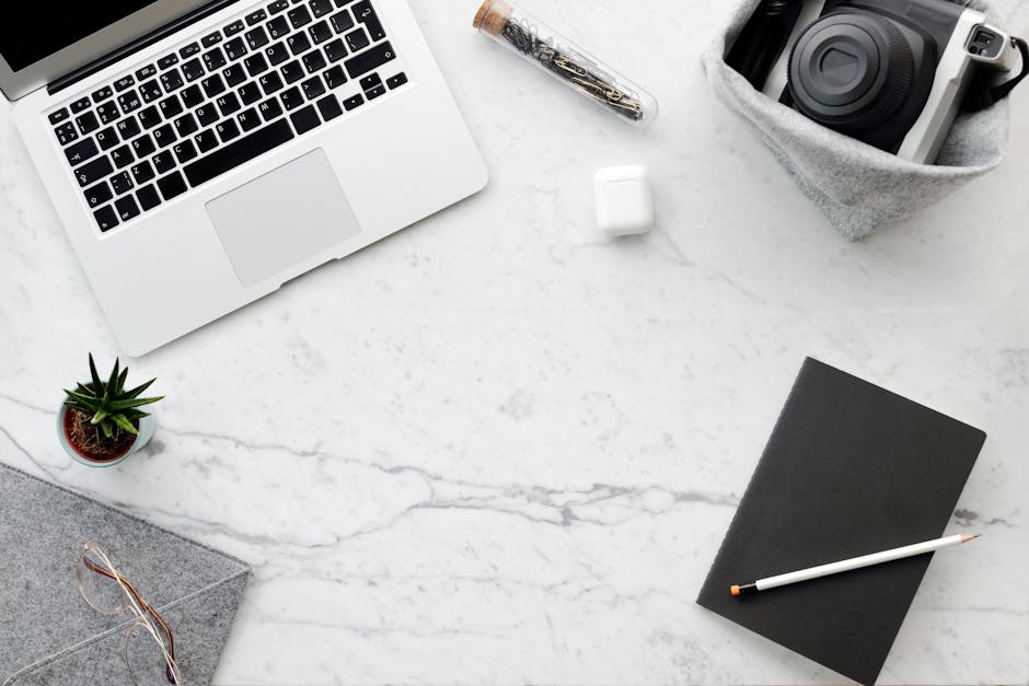 Top view of a modern workspace on a marble desk featuring a laptop, camera, plant, and stationery.